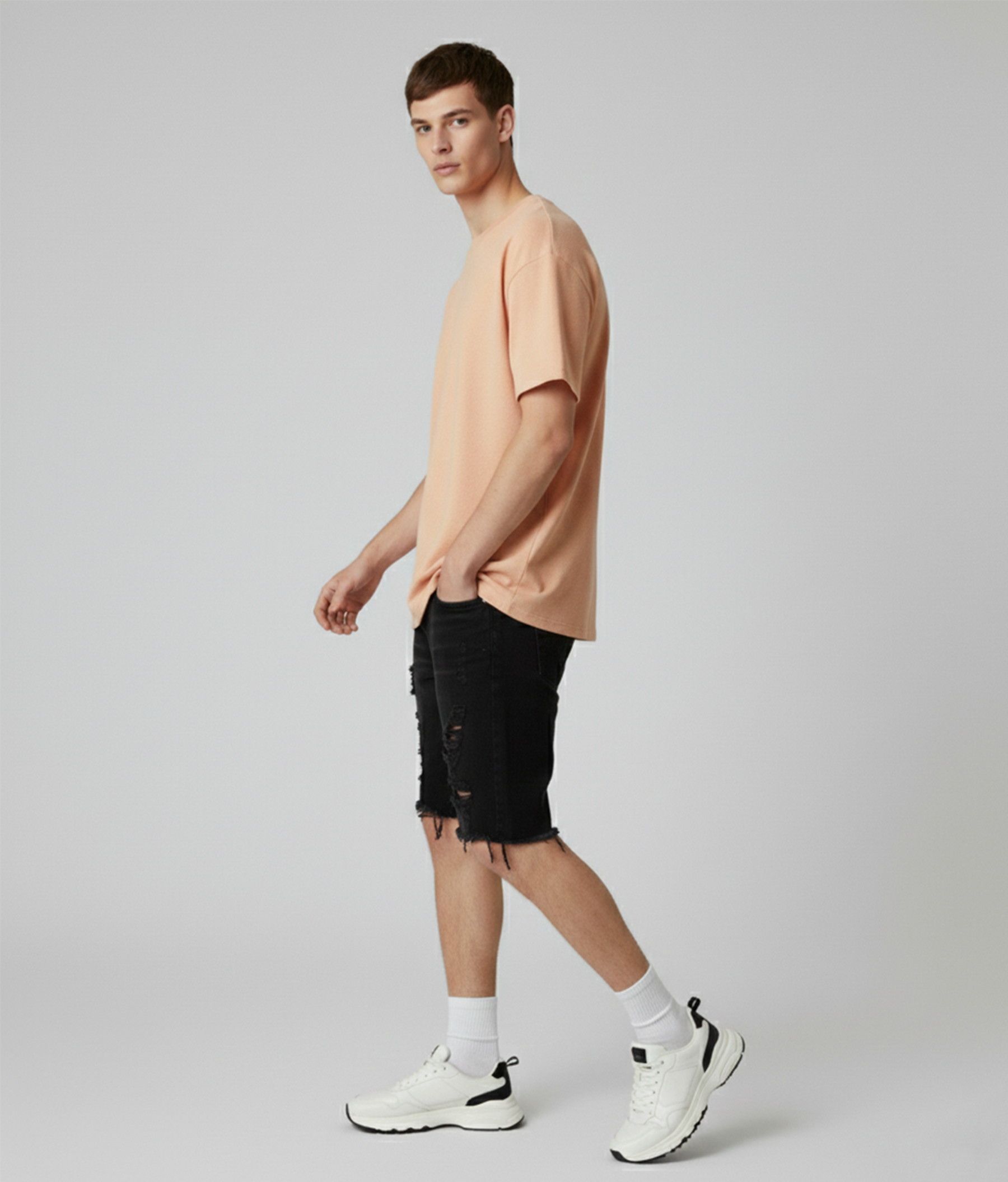 Man wearing a beige t-shirt, black shorts, and white sneakers on a plain background